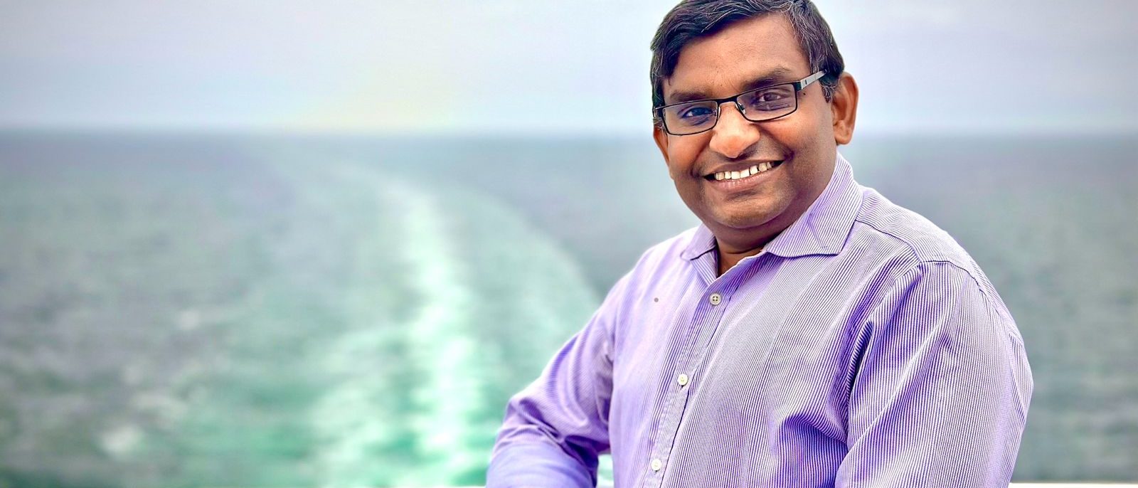 Bejoy Mathew smiling, leaning on a railing with the sea behind him